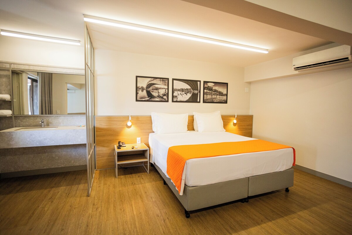 A hotel room with a bed, bedside table, and a mirror. There are three framed pictures on the wall.