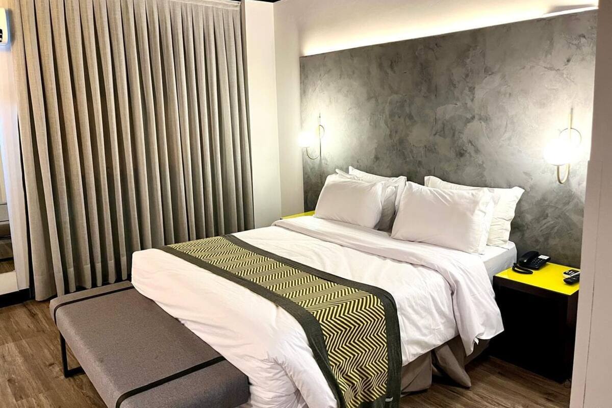 Una camera d'albergo con un letto, un comodino, una lampada e una panca.