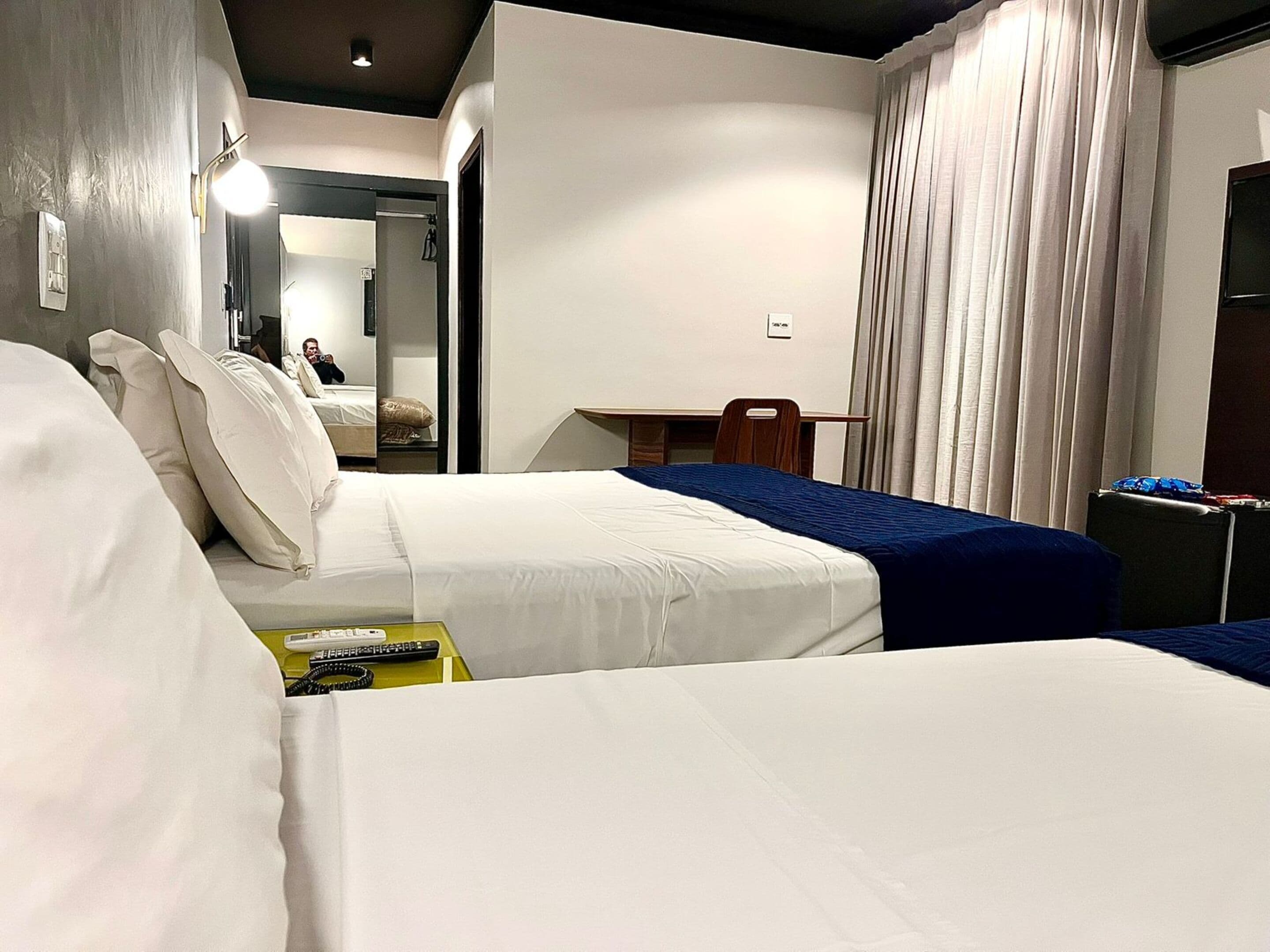 A hotel room with two beds, a desk, a chair, a mirror, and a window with curtains.