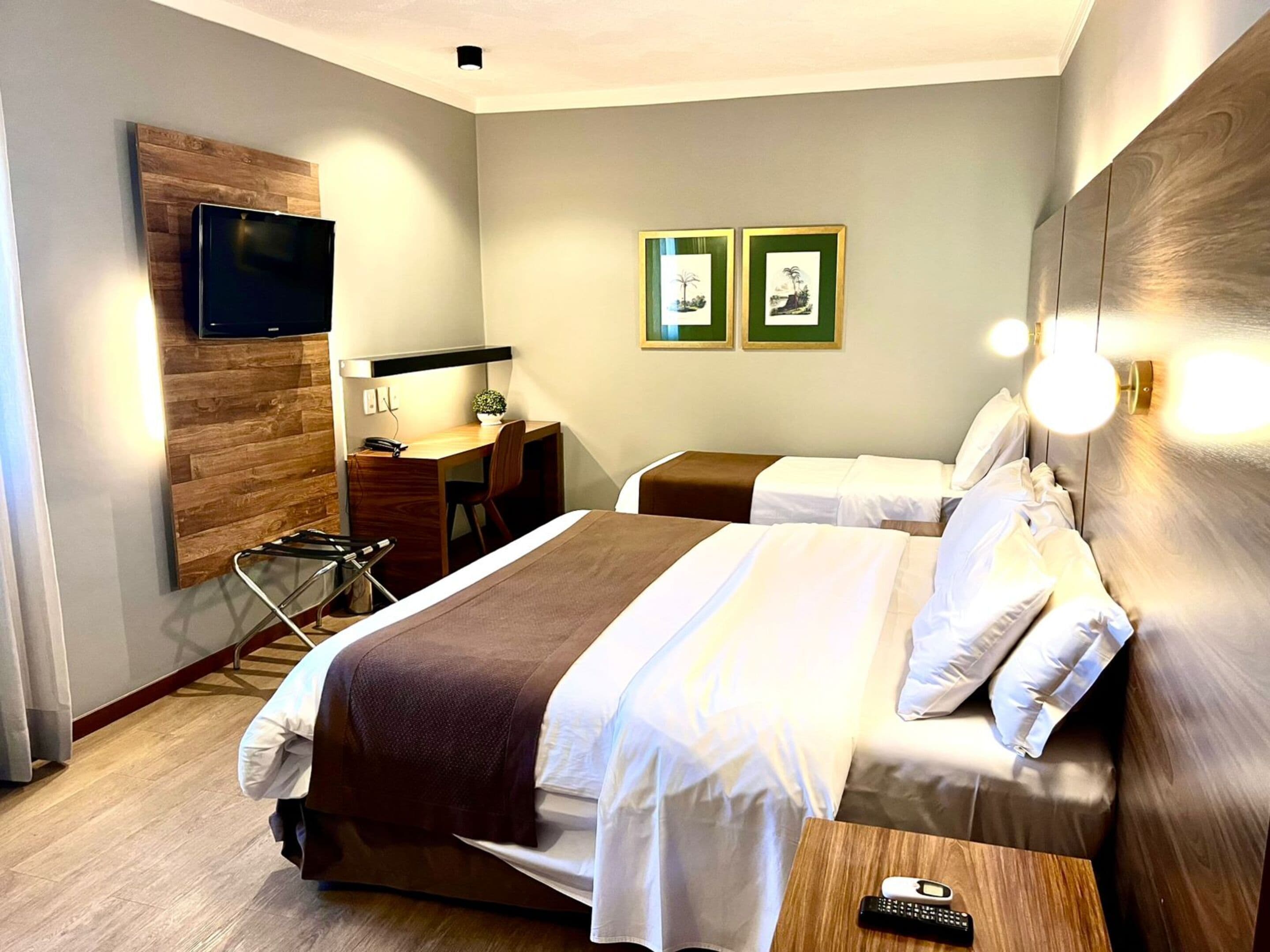 A hotel room with two beds, a desk, a chair, a TV mounted on a wooden panel, and two framed pictures on the wall.