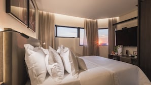 A hotel room with a large bed, a flat-screen TV, a desk with a lamp, and a window with curtains.