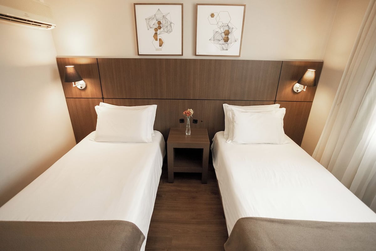 Une chambre d’hôtel avec deux lits simples, une tête de lit en bois, des lampes fixées au mur et deux œuvres d’art abstraites encadrées.