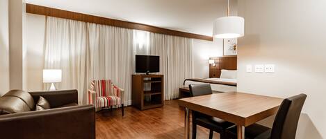 A modern hotel room with a wooden dining table, leather sofa, and a flat-screen TV.