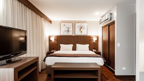 Une chambre d’hôtel dotée d’un grand téléviseur à écran plat, d’un cadre de lit en bois, d’une table de chevet et d’un banc. Deux appliques murales et des œuvres d’art sont disposées au-dessus du lit.