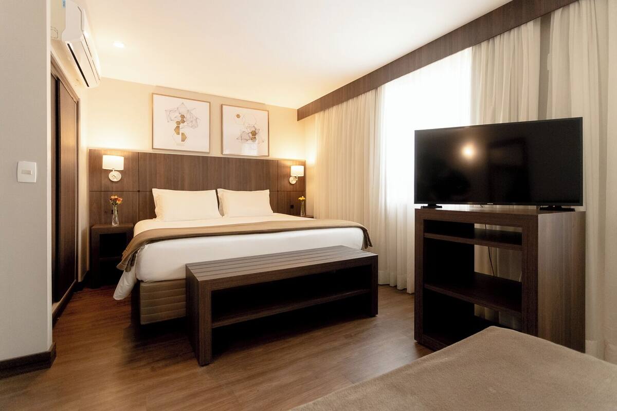 Une chambre d’hôtel moderne avec un grand lit, des lampes de chevet, un téléviseur à écran plat et un banc en bois.