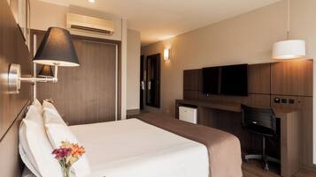 Quarto de hotel com cama, luminária de cabeceira, TV e cozinha compacta.