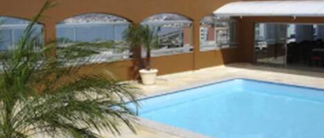 A swimming pool with a tiled deck, palm trees, and a building with large windows and a covered area.