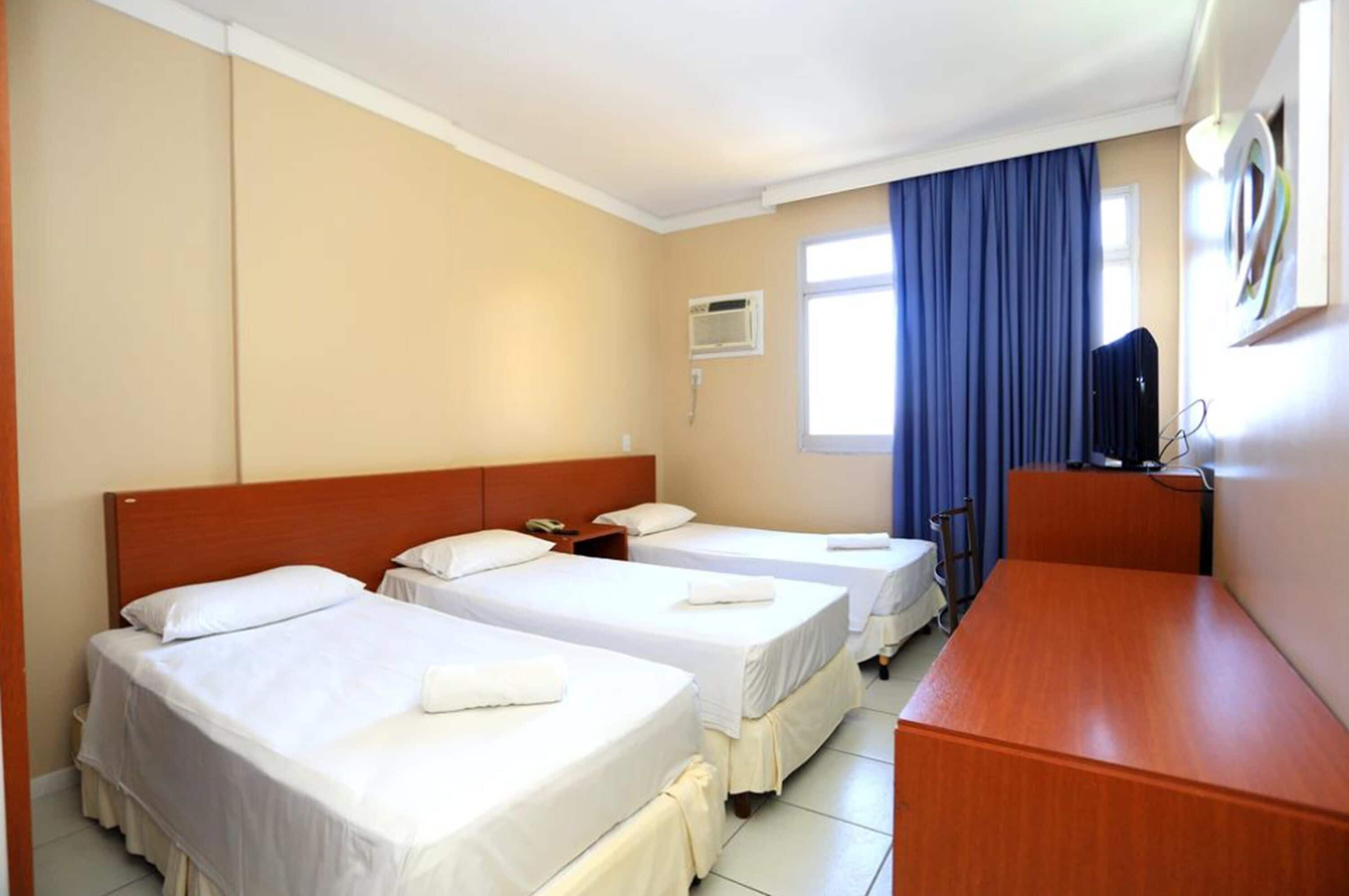 Habitación de hotel con dos camas individuales, cabecero de madera, escritorio y ventana con cortinas azules.