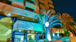 Ett hotell med modern design, med en imponerande trappa som leder till entrén, ett stort palmträd framför och en skylt som visar hotellets namn.