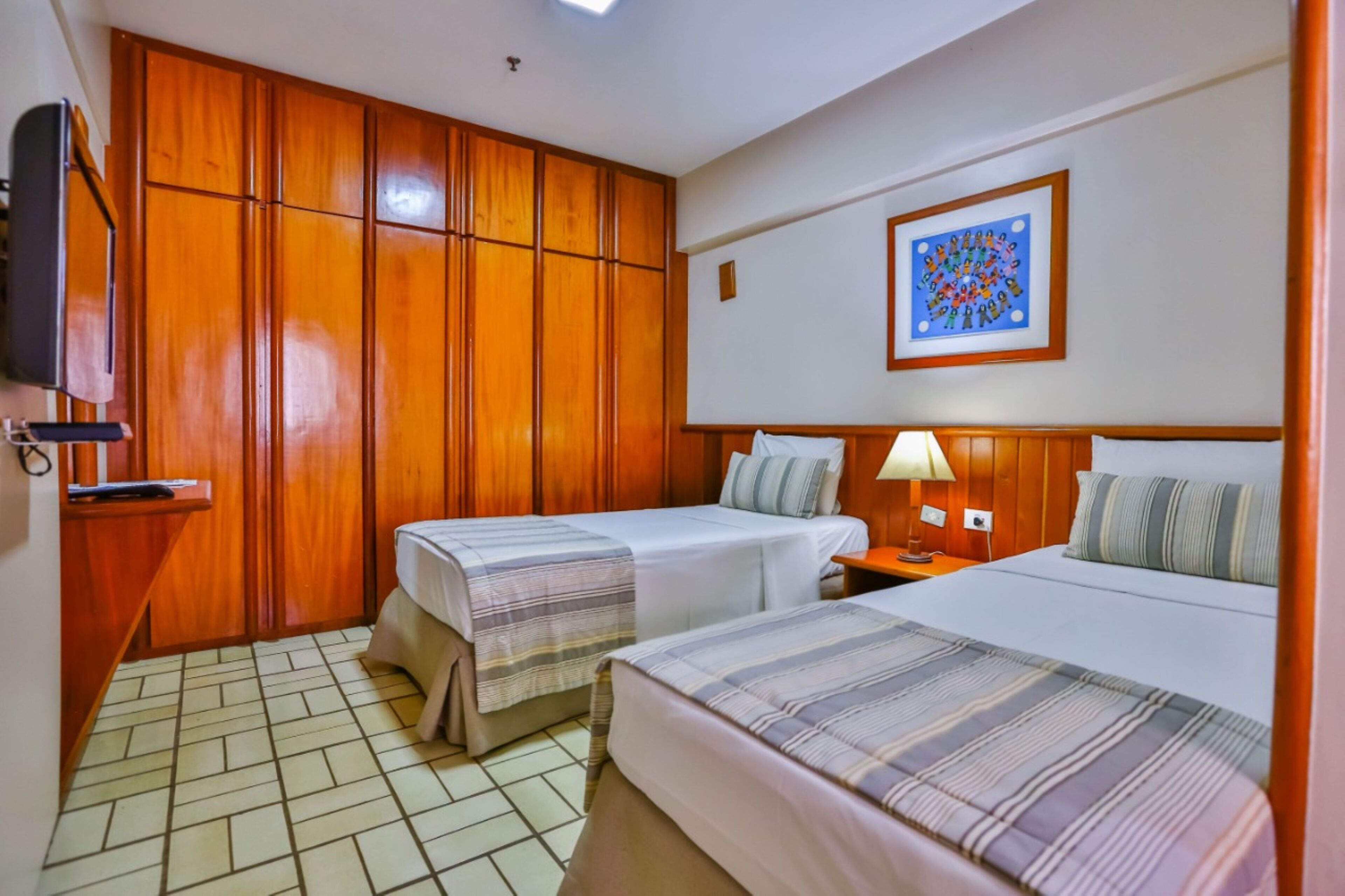 A hotel room with two single beds, a wooden wardrobe, a television, and a framed picture on the wall.