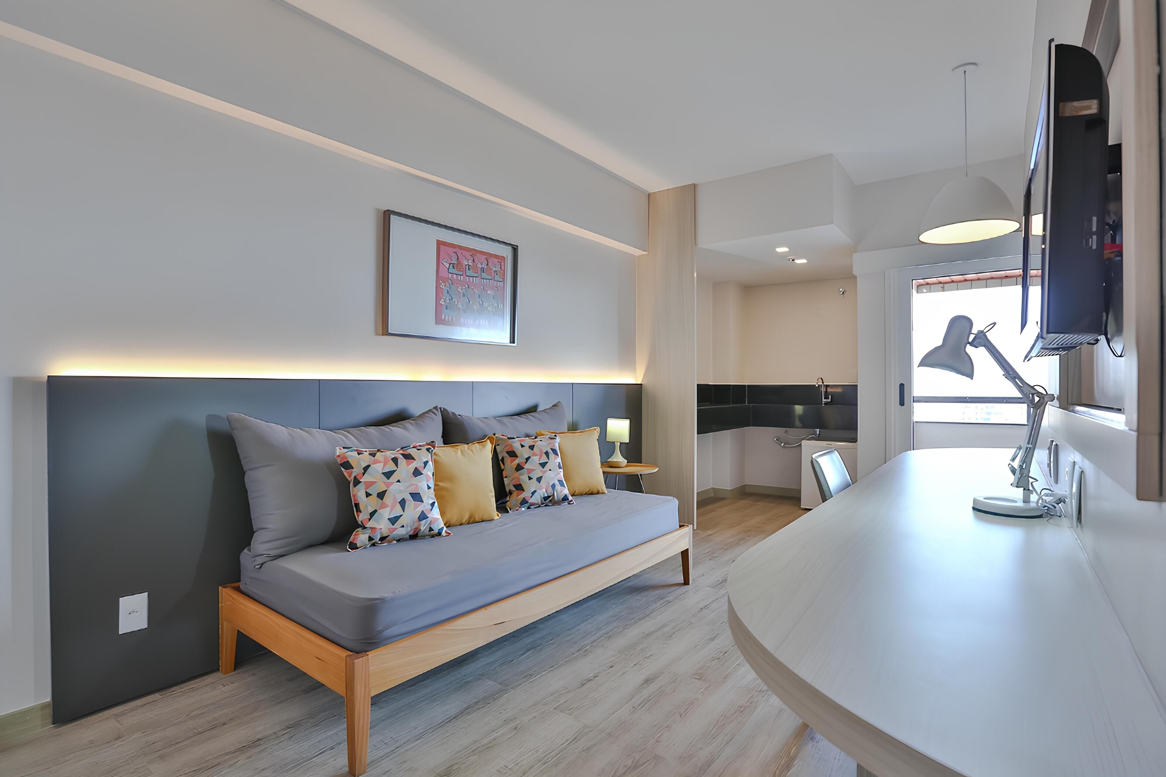 Un salon moderne avec un canapé, un téléviseur fixé au mur et un coin cuisine visible à travers une porte ouverte.