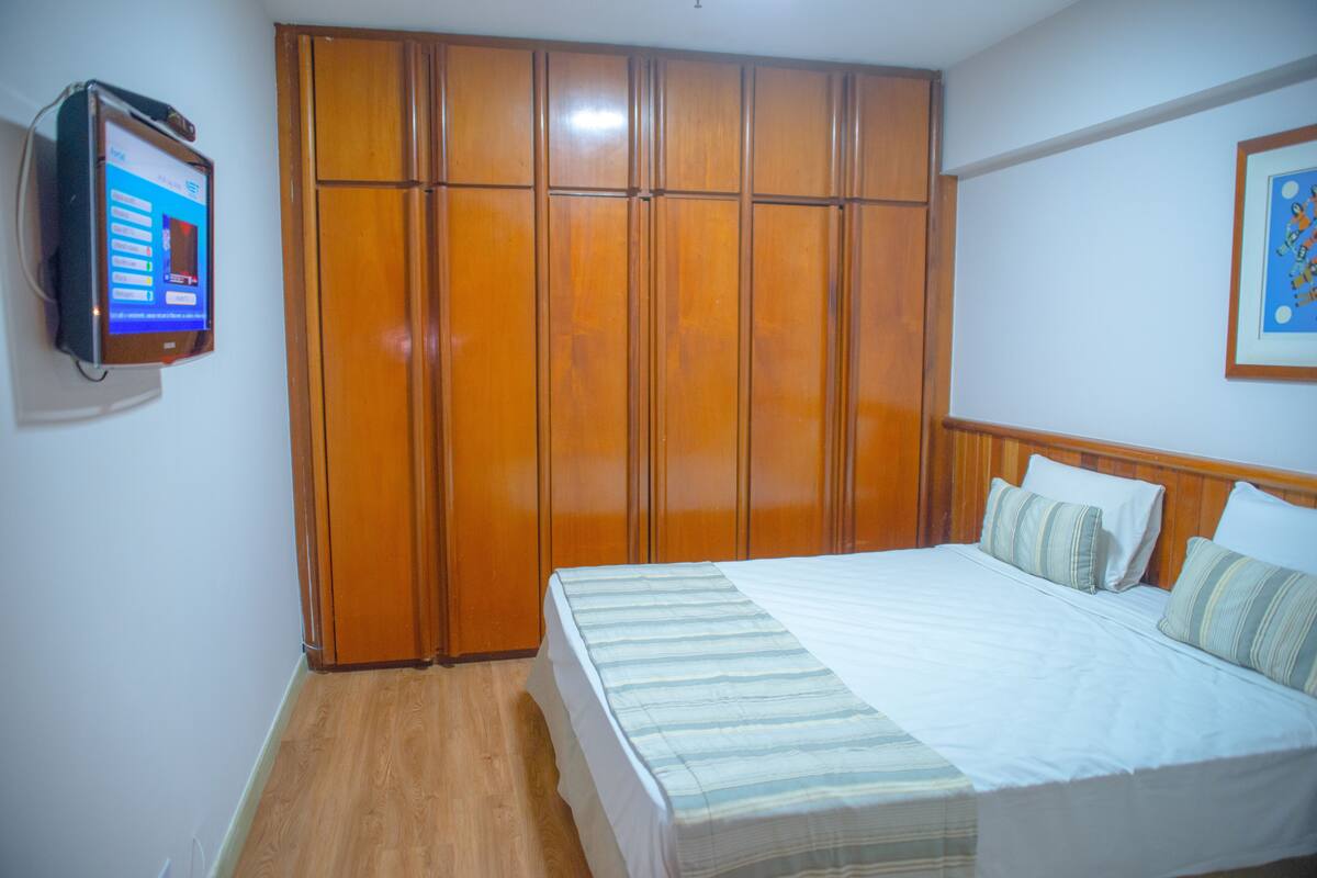 Une chambre avec une armoire en bois, un lit avec une literie à rayures et un téléviseur fixé au mur.