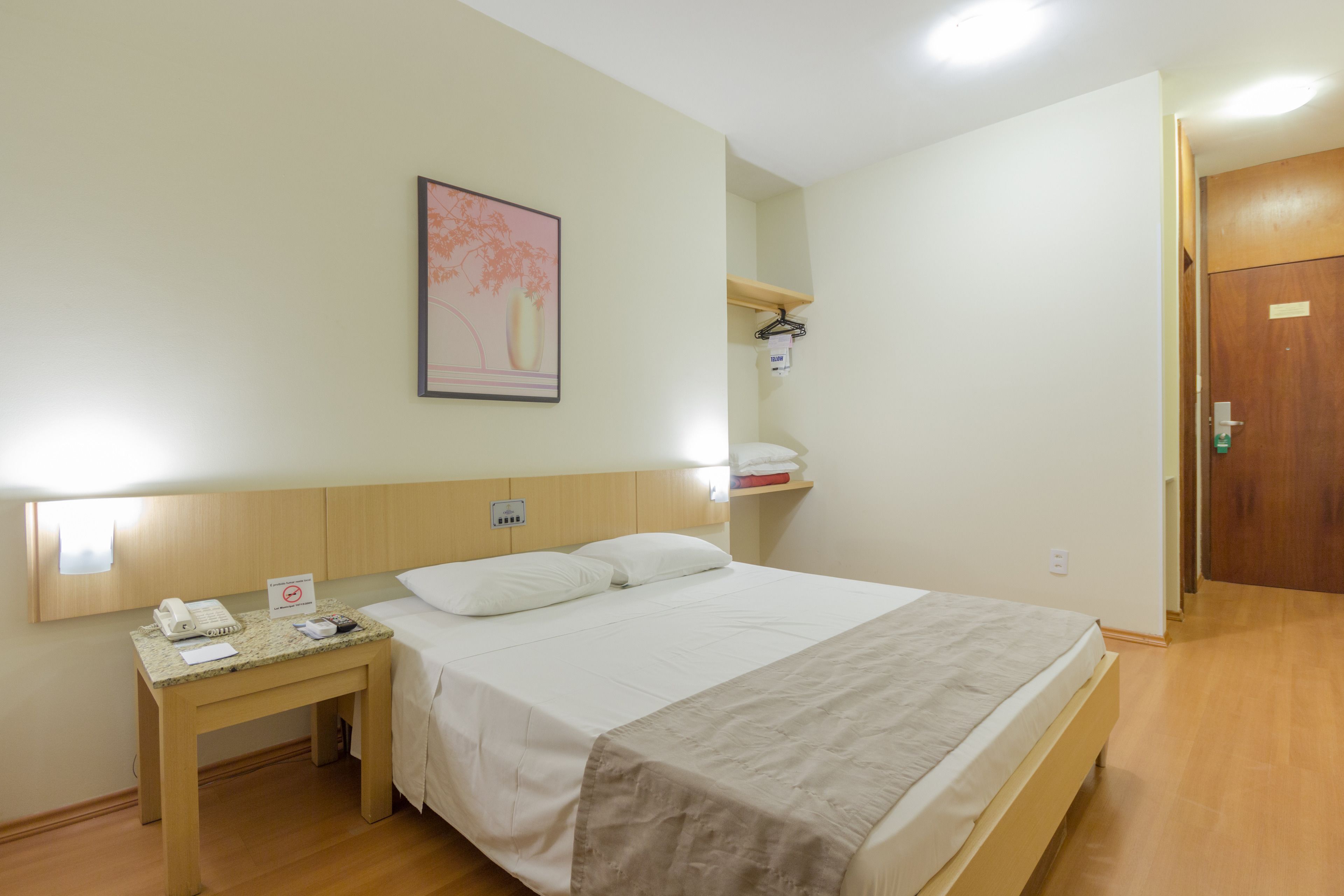 A hotel room with a bed, bedside table, and a wall-mounted shelf with a towel rack.