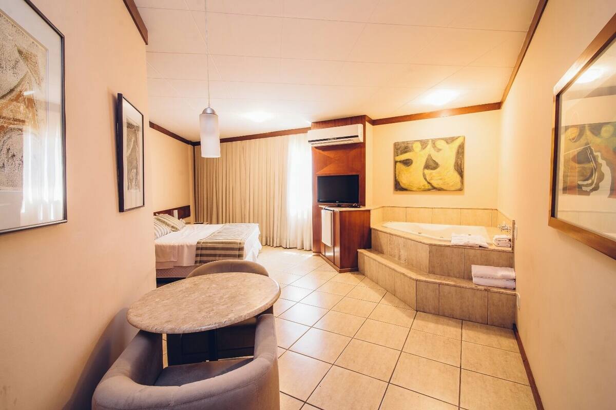 Ett hotellrum med en säng, ett litet bord, en soffa, en TV och ett badrum med badkar, samt konstverk på väggarna.