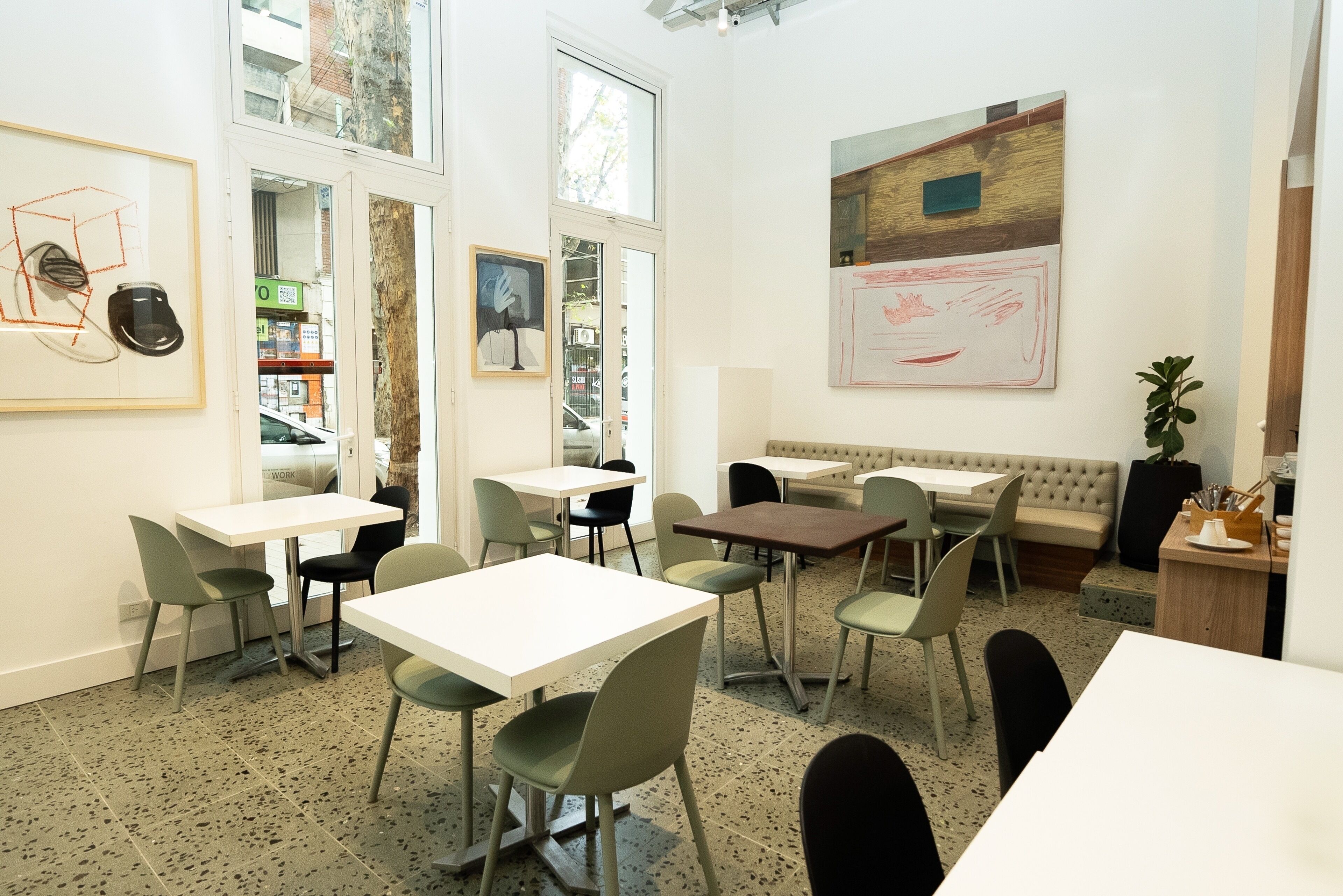 A modern dining area with white tables and green chairs, large windows, and artwork on the walls.