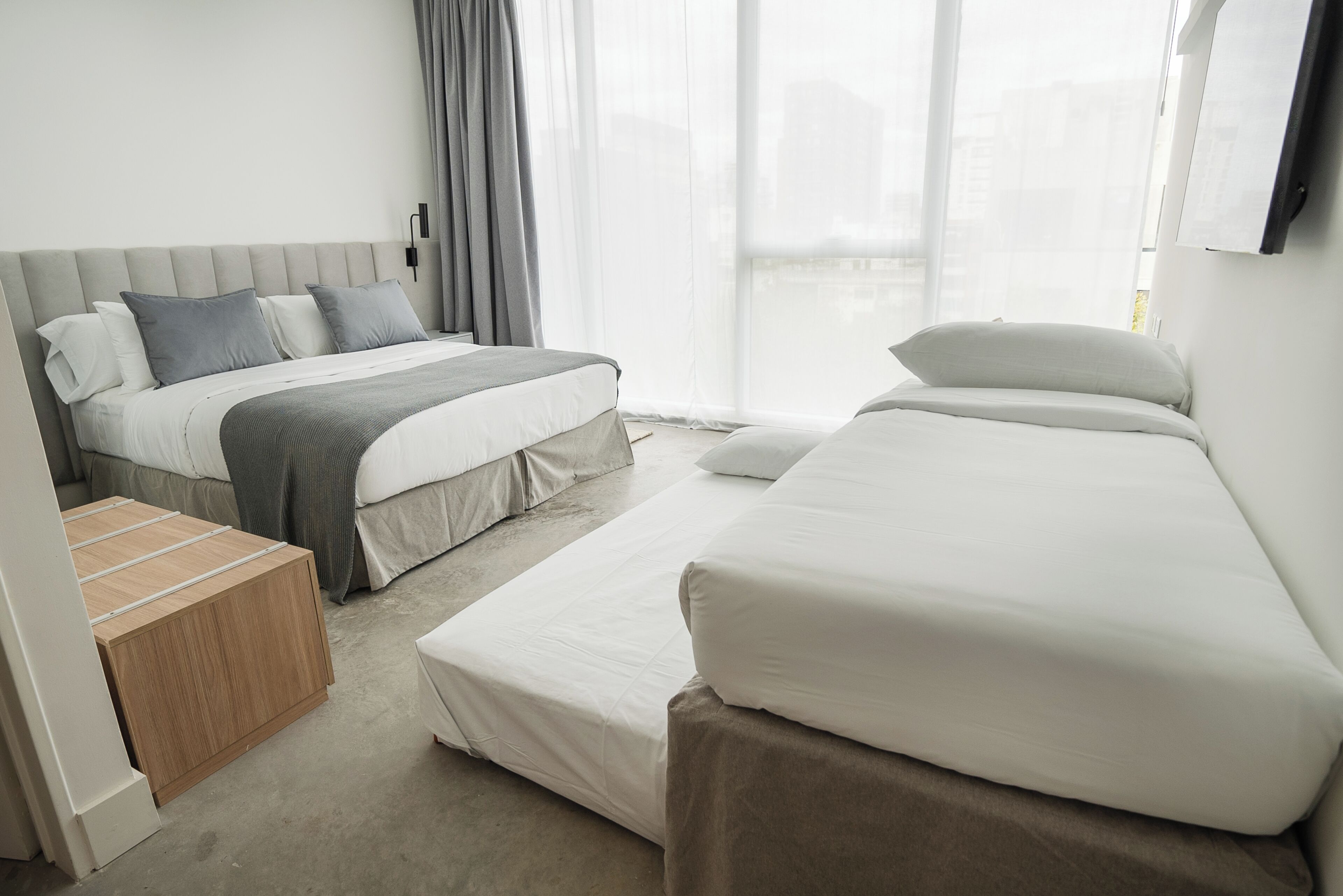 Habitación de hotel moderna con una cama grande, una cama más pequeña, una mesita de noche de madera y vistas de la ciudad a través de amplias ventanas.