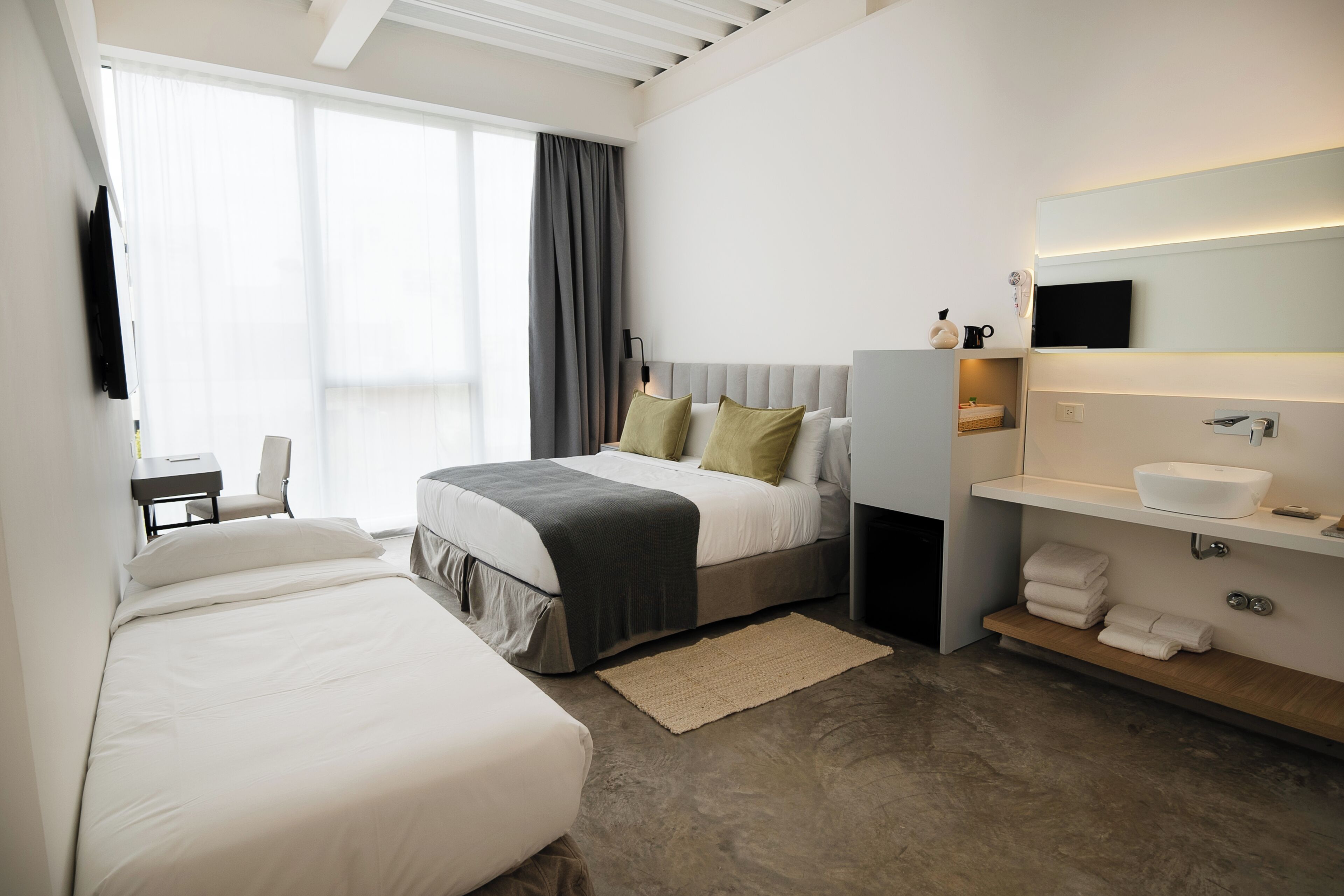 Una habitación de hotel moderna con una cama grande, una cama más pequeña, un escritorio y un baño con espejo y lavamanos.