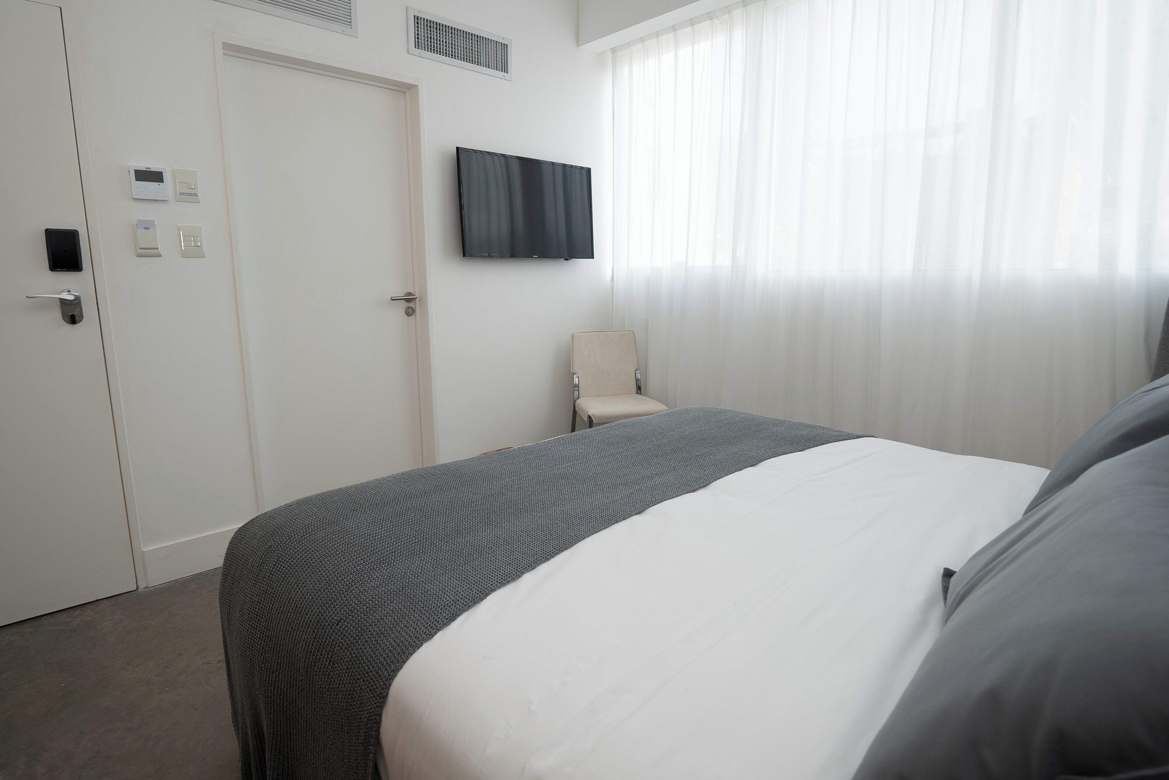 Une chambre d’hôtel comprenant un lit, une chaise, un téléviseur fixé au mur, une porte dotée d’une serrure et d’un panneau de commande.