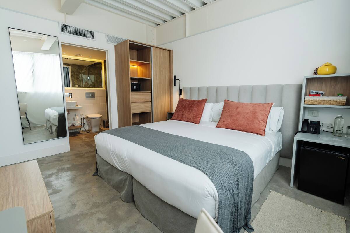 A modern hotel room with a large bed, a wardrobe, a small desk, and a mini-fridge.