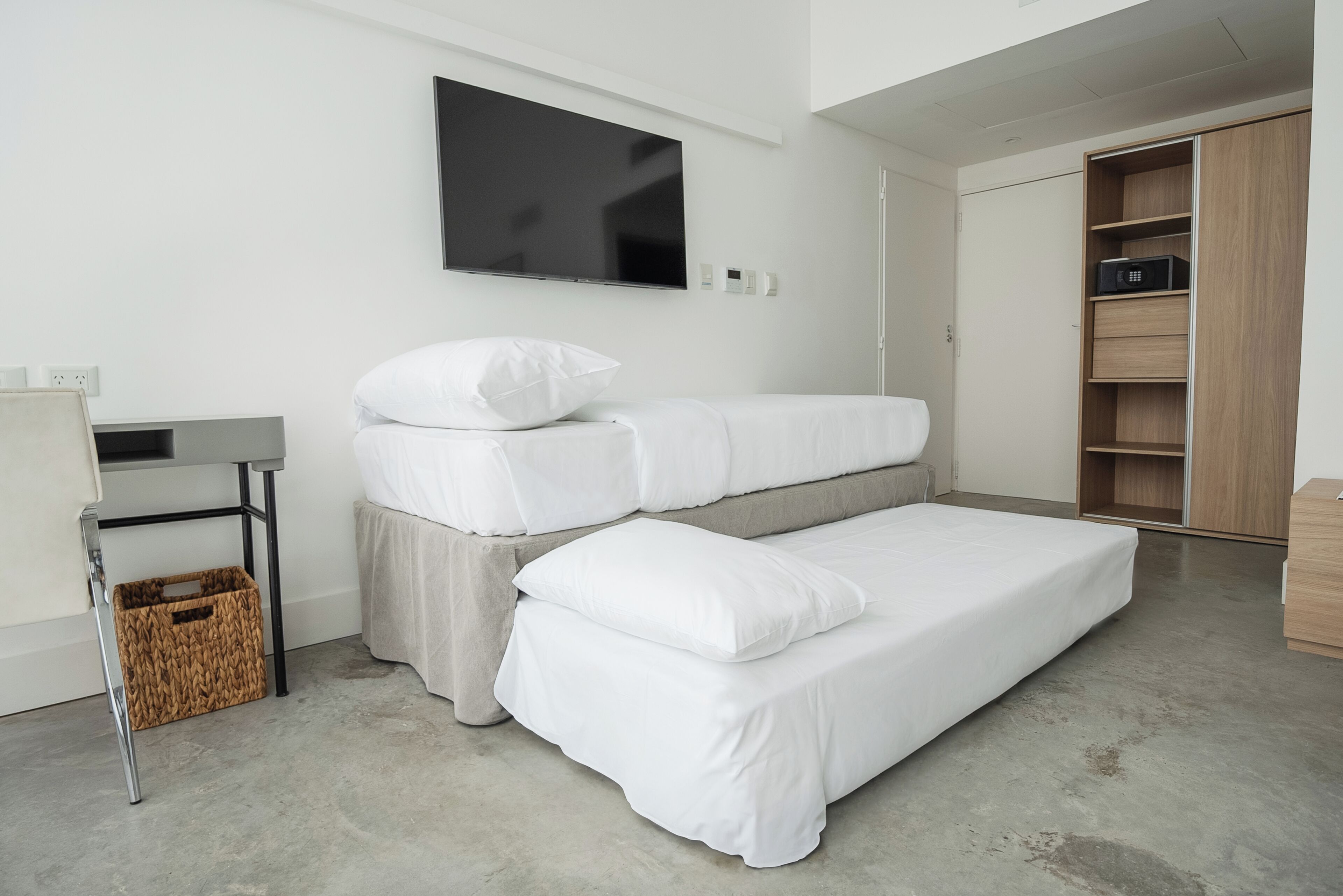 A modern bedroom with a bed, a TV mounted on the wall, a wooden wardrobe, and a small table with a basket underneath.