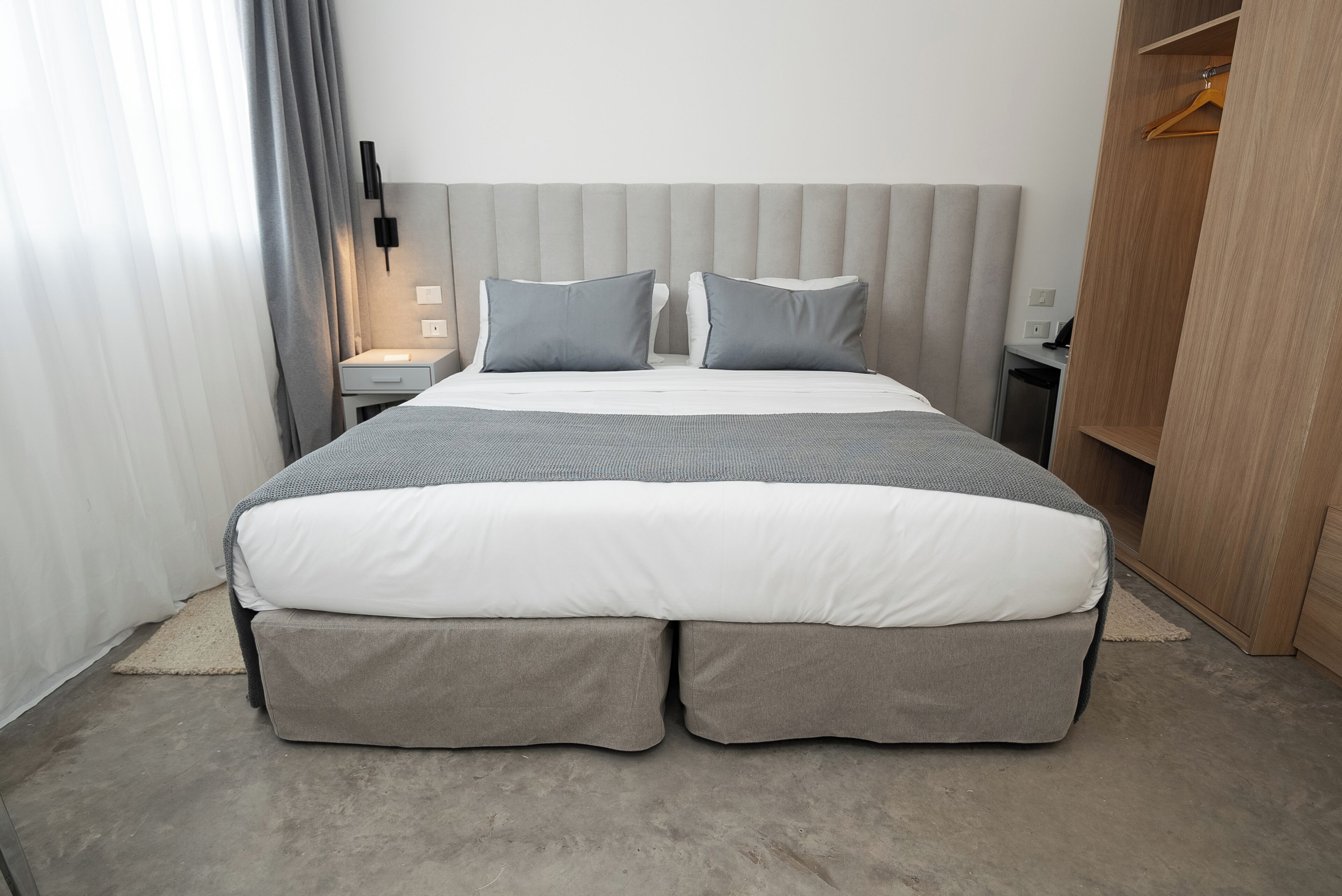 Een moderne hotelkamer met een groot bed, grijs beddengoed, een nachtkastje met een lamp en een houten garderobe.