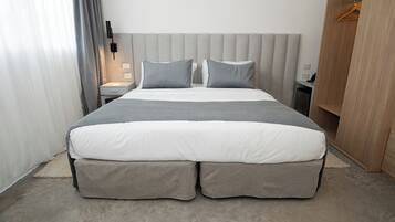 Een moderne hotelkamer met een groot bed, grijs beddengoed, een nachtkastje met een lamp en een houten garderobe.