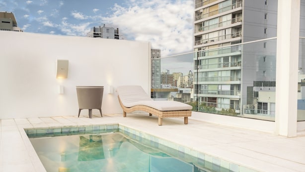 A rooftop pool area with a lounge chair and a small table, surrounded by a white wall and a glass railing overlooking a cityscape. - Vain Boutique Hotel (Buenos Aires)