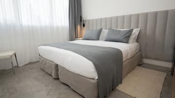 A modern bedroom with a large bed, grey bedding, a bedside table, and a window with curtains.
