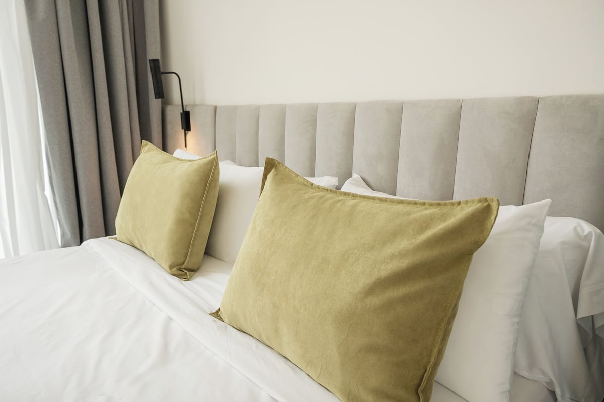 A neatly made bed with white linens and two green pillows, a grey upholstered headboard, and a black wall-mounted lamp.