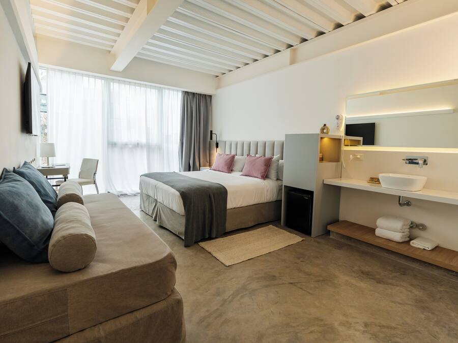 Una habitación de hotel moderna con cama, sofá, mesita de noche, lámpara, espejo y zona de baño con lavabo y perchero para toallas.