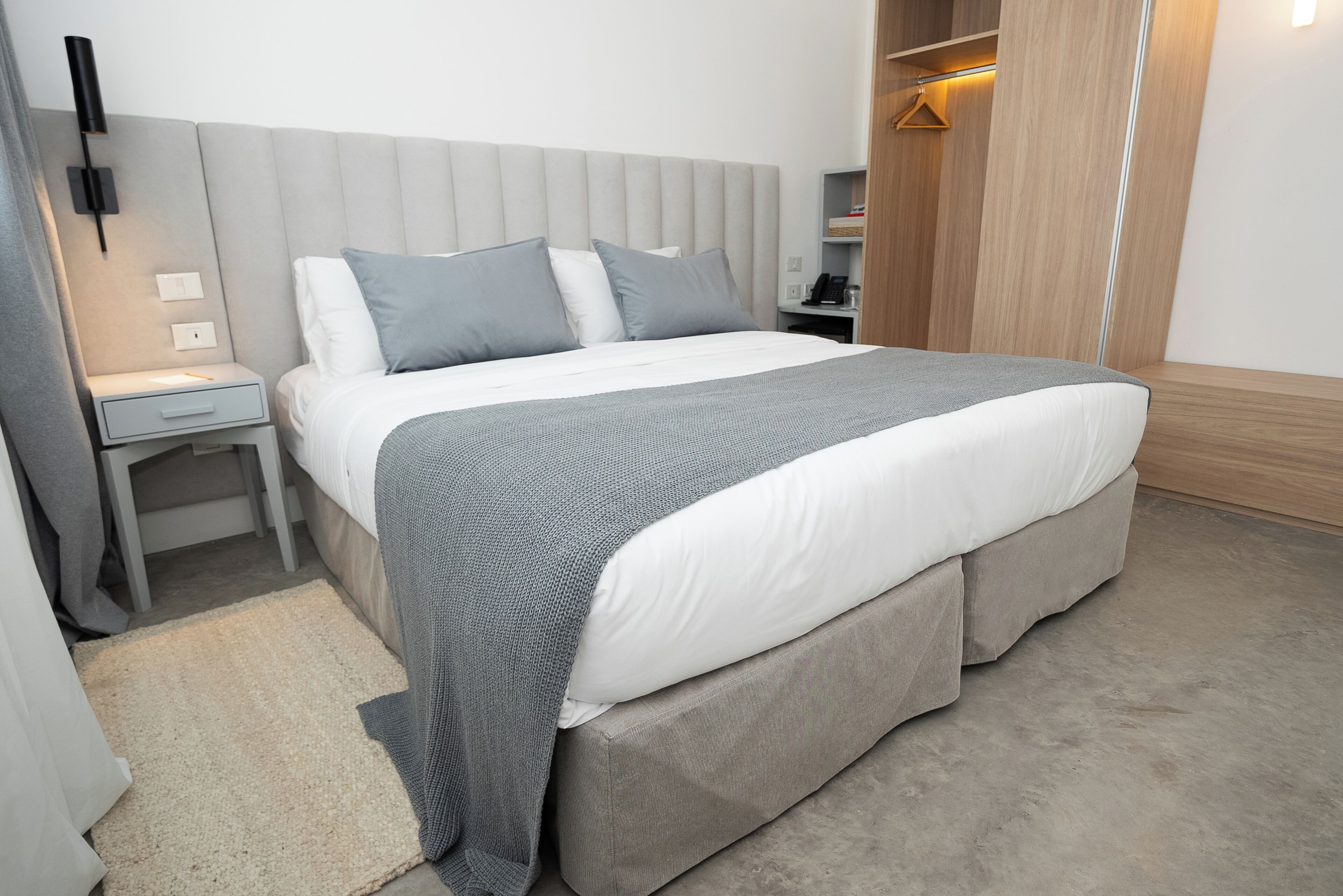 A modern hotel room with a large bed, a bedside table, a nightstand, a wardrobe, and a small seating area.