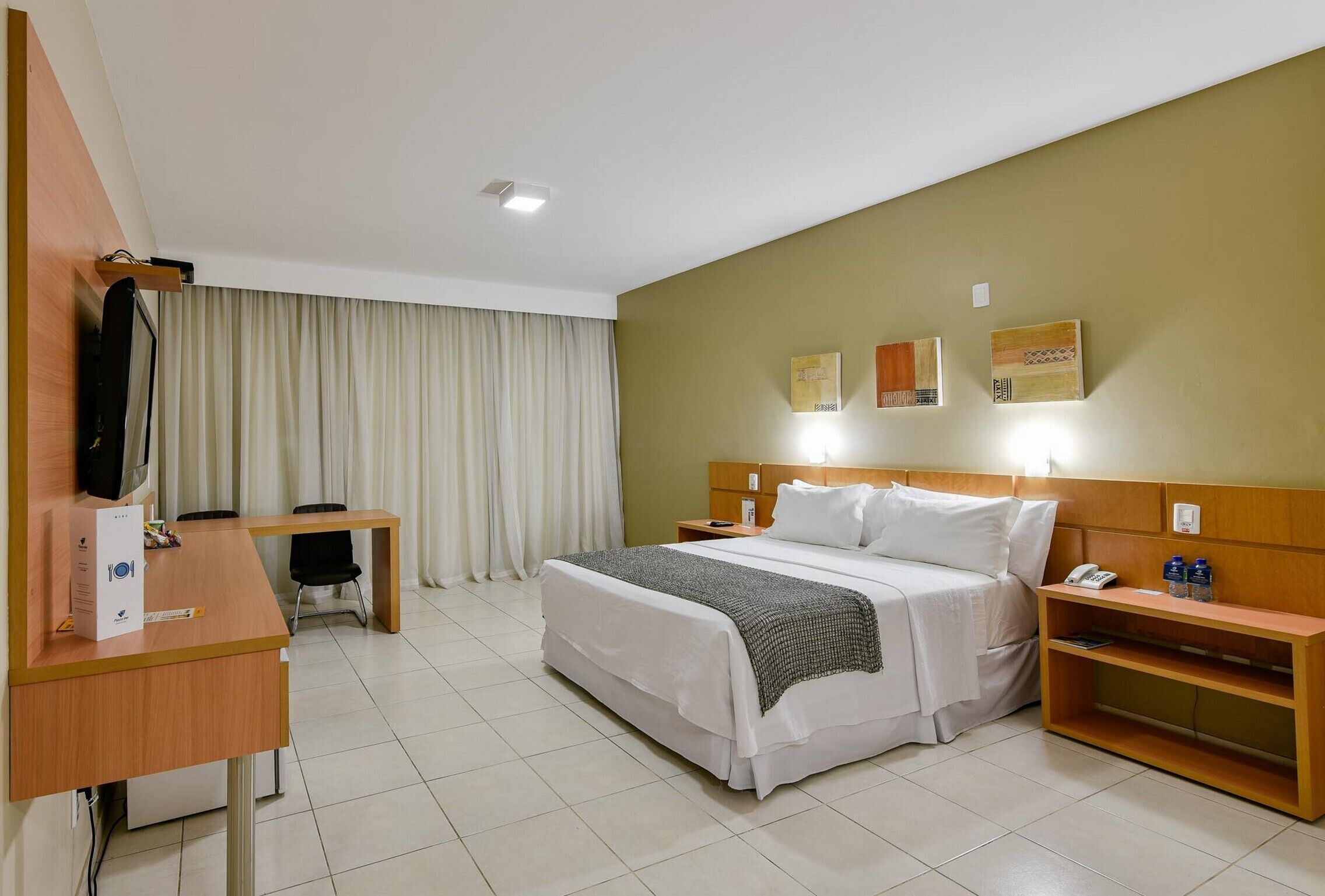 Habitación de hotel con cama, mesita de noche, escritorio, silla, cortinas y una obra de arte en la pared.