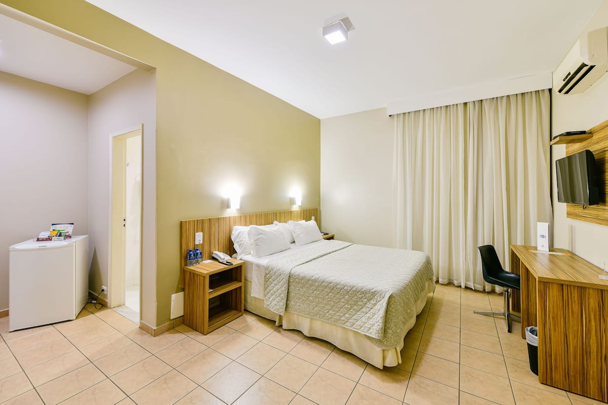 A hotel room with a bed, bedside tables, a desk, a chair, a TV, and an air conditioning unit.