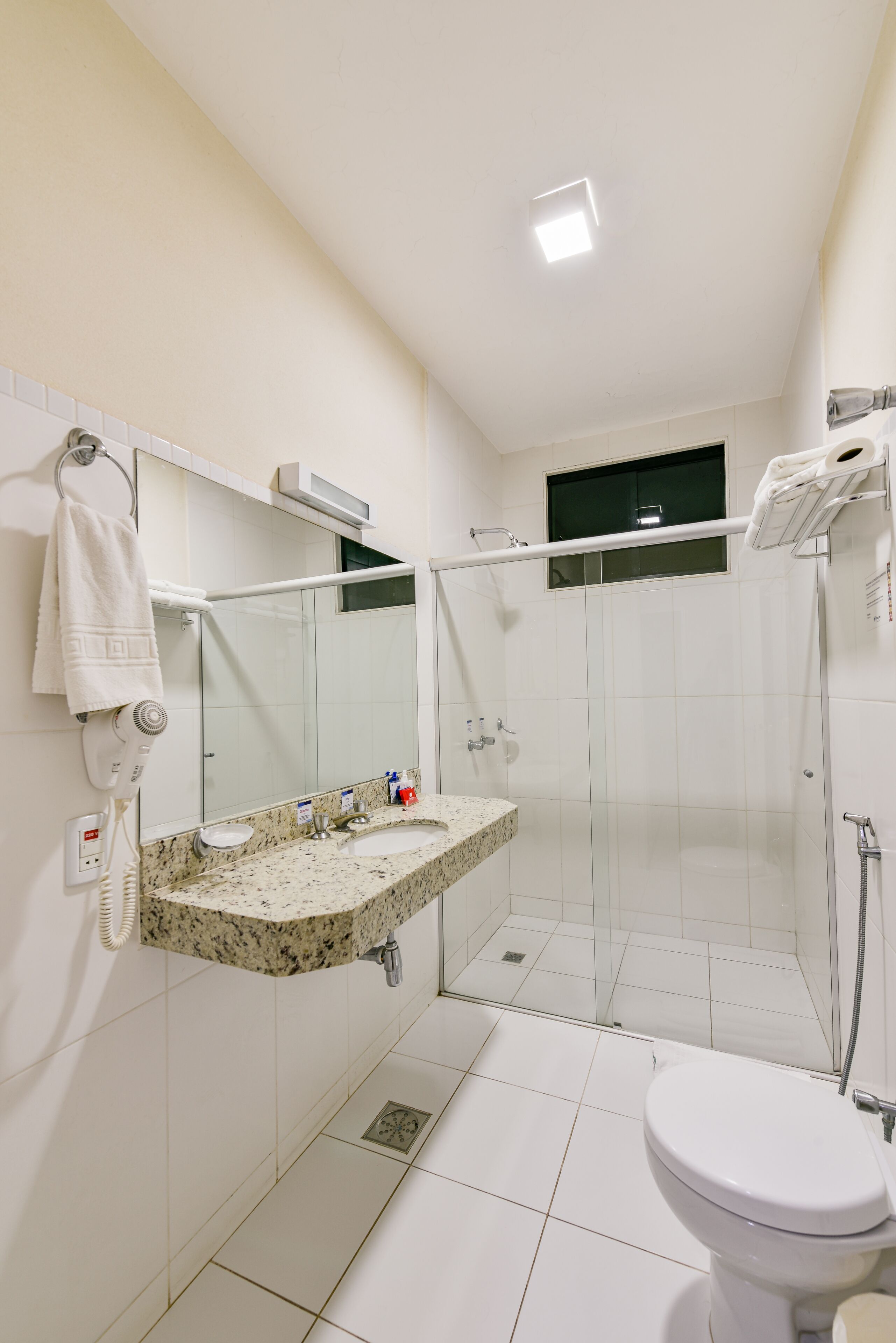 A modern bathroom with a white sink, mirror, and toilet. It includes a glass-enclosed shower area and a hairdryer on the wall.