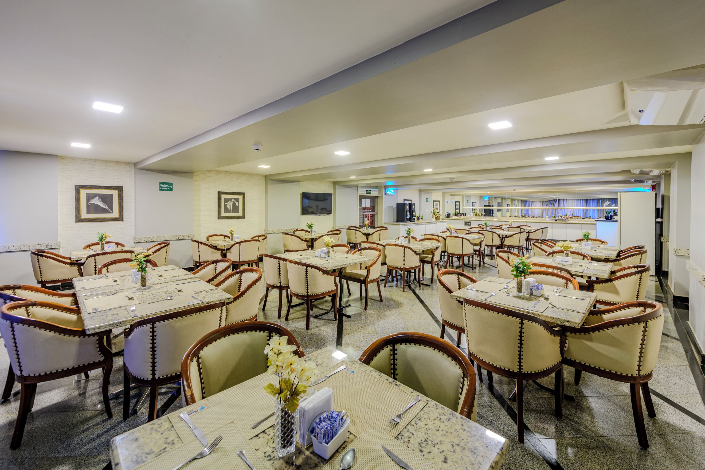 Uma sala de jantar de hotel com várias mesas e cadeiras, com louças postas e centros de mesa florais.