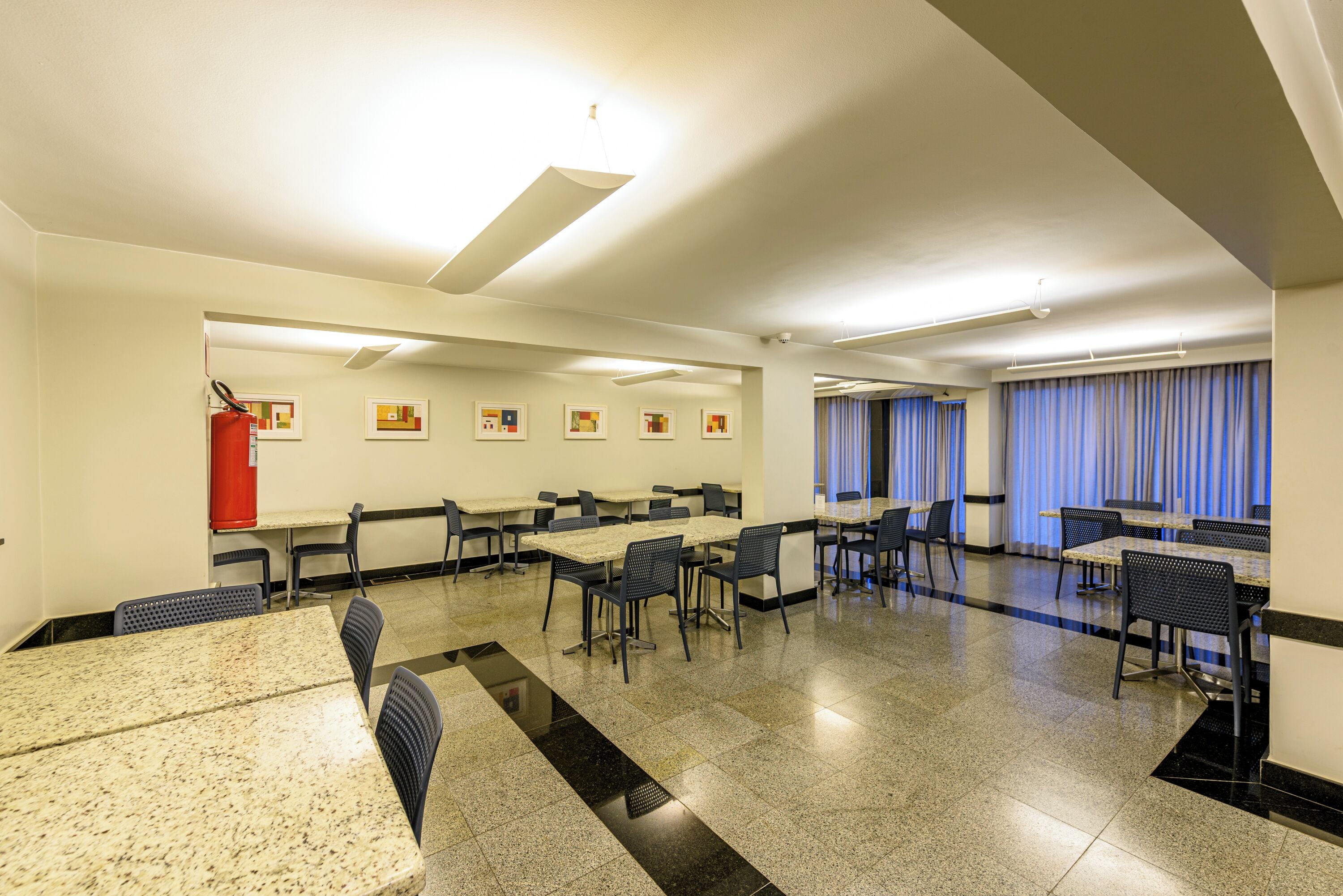 A hotel dining area with multiple tables and chairs, ceiling lights, and artwork on the walls.