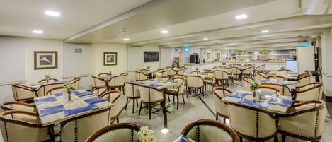 Comedor de hotel con varias mesas y sillas, una cocina al fondo, y un ambiente limpio y bien iluminado.