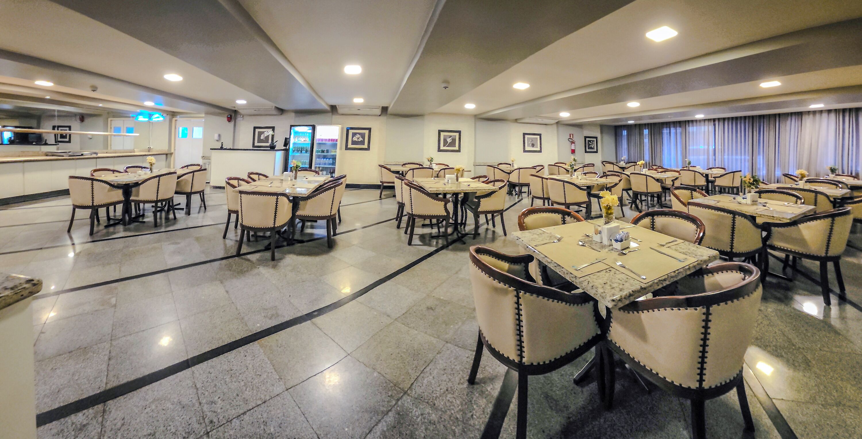 Un comedor de hotel con varias mesas y sillas, una cocina al fondo y cuadros en las paredes.