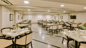A hotel dining area with multiple tables and chairs, arranged in rows, with ceiling lights and framed pictures on the walls. - Plaza Inn Augustus - Downtown (Goiania)