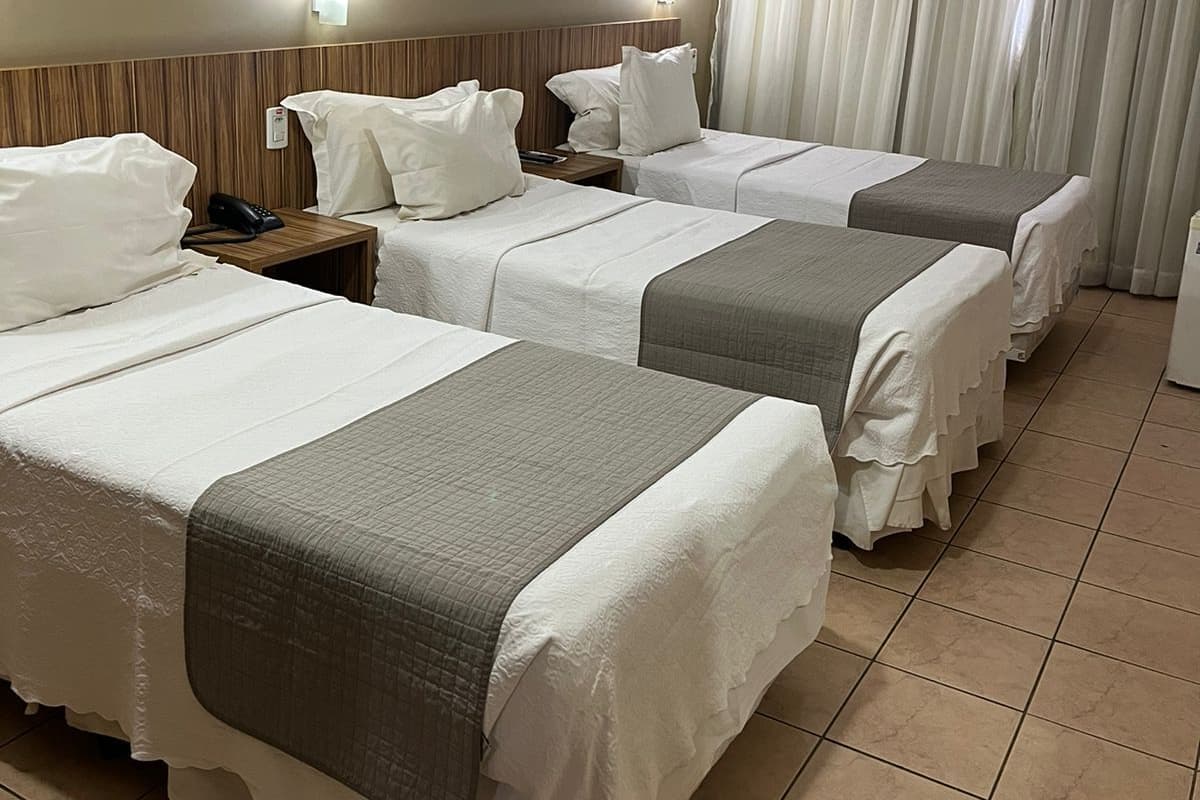 A hotel room with four beds, each with white bedding and a grey blanket. There are two bedside tables with lamps, a window with curtains, and a tiled floor.