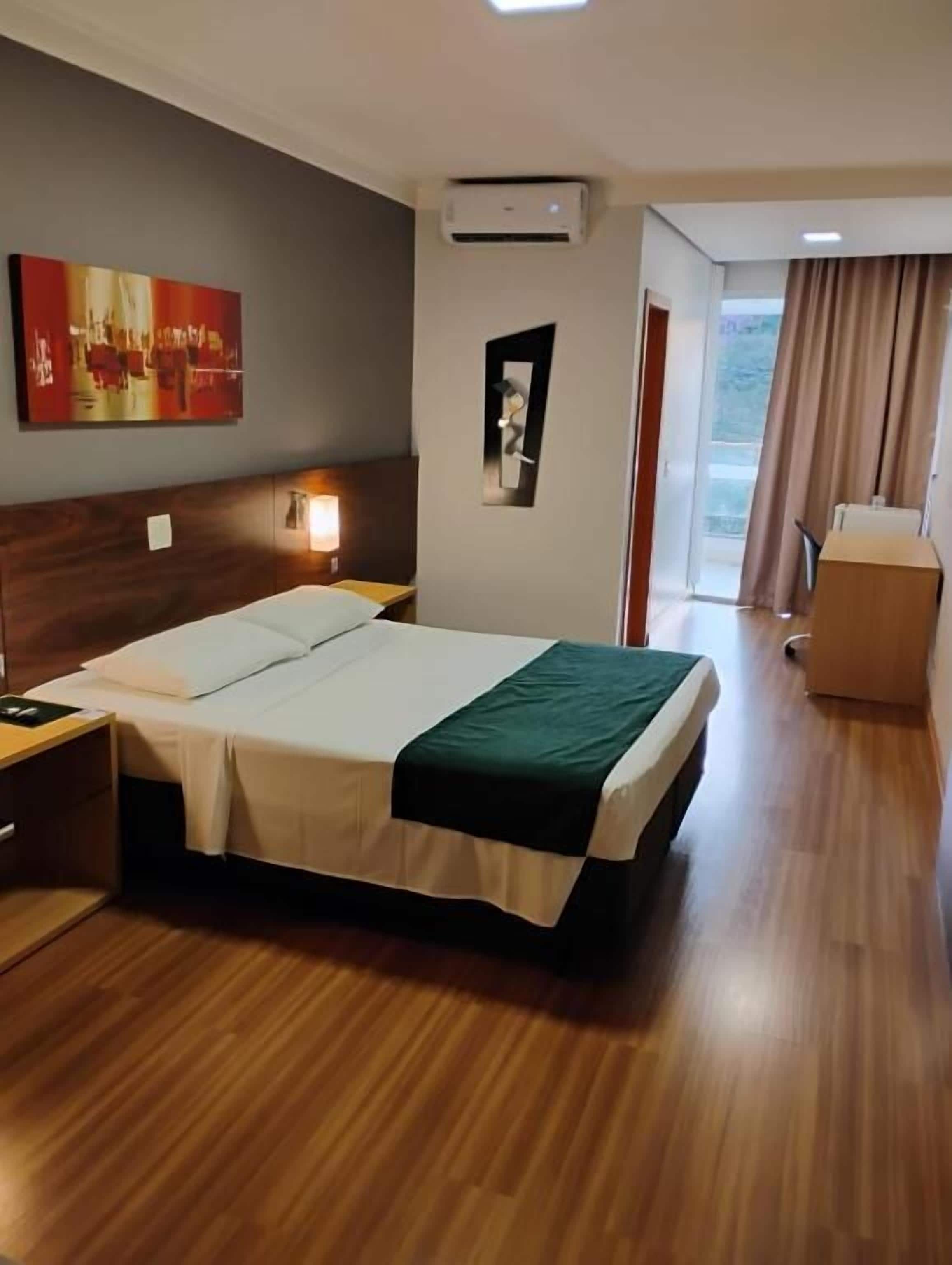 A hotel room with a wooden floor, a bed with white linens, a nightstand, a wall-mounted air conditioning unit, a painting, and a view through a window with curtains.