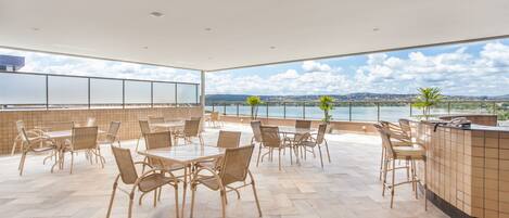 A spacious outdoor dining area with wicker chairs and a tiled floor, overlooking a body of water and distant landscape.
