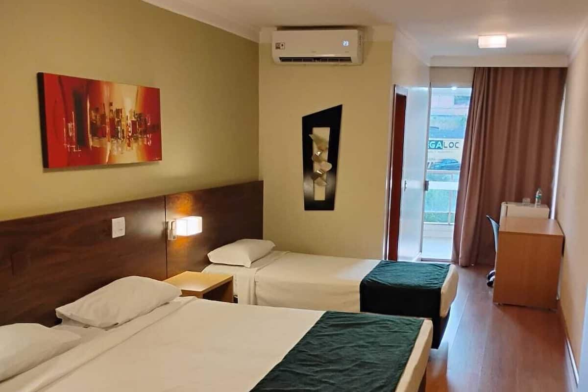 A hotel room with two beds, a ceiling fan, an air conditioning unit, a painting on the wall, and a view of the outside through a window.