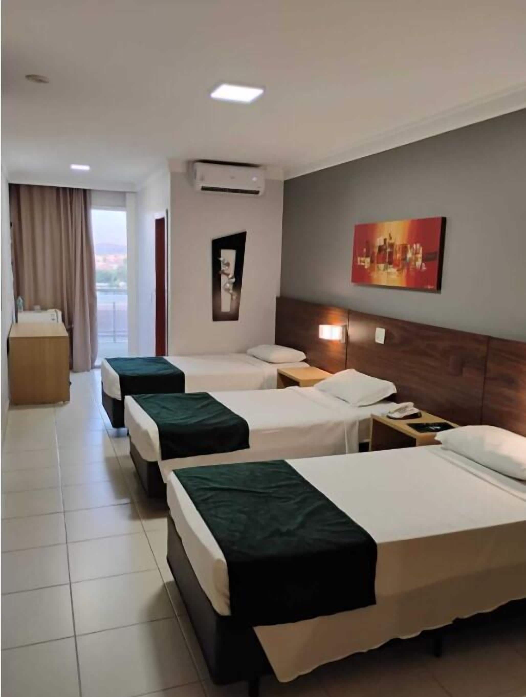 A hotel room with three single beds, a wooden headboard, a bedside table with a lamp, an air conditioning unit, a painting on the wall, and a view of the outside.