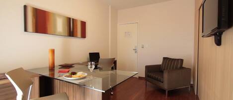 A modern living room with a glass dining table, two chairs, a sofa, and a flat-screen TV mounted on the wall.