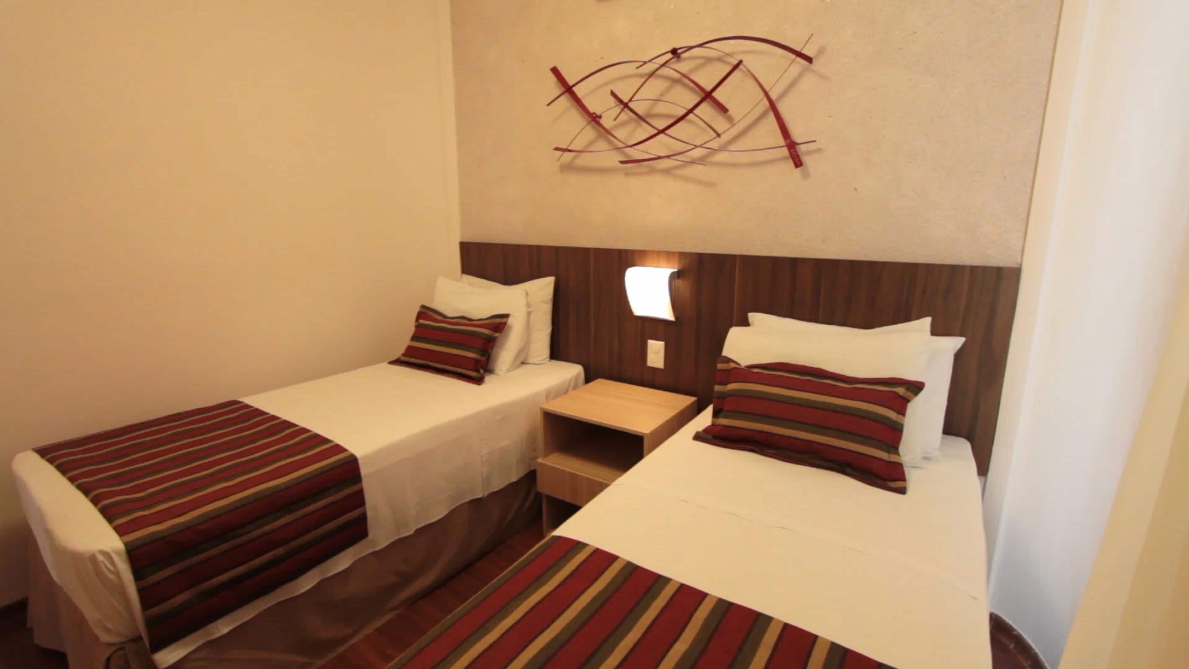 Um quarto pequeno e acolhedor com duas camas de solteiro, cada uma com roupa de cama e almofadas listradas. Há uma mesa de cabeceira com uma luminária entre as camas, e uma cabeceira de madeira acima delas.
