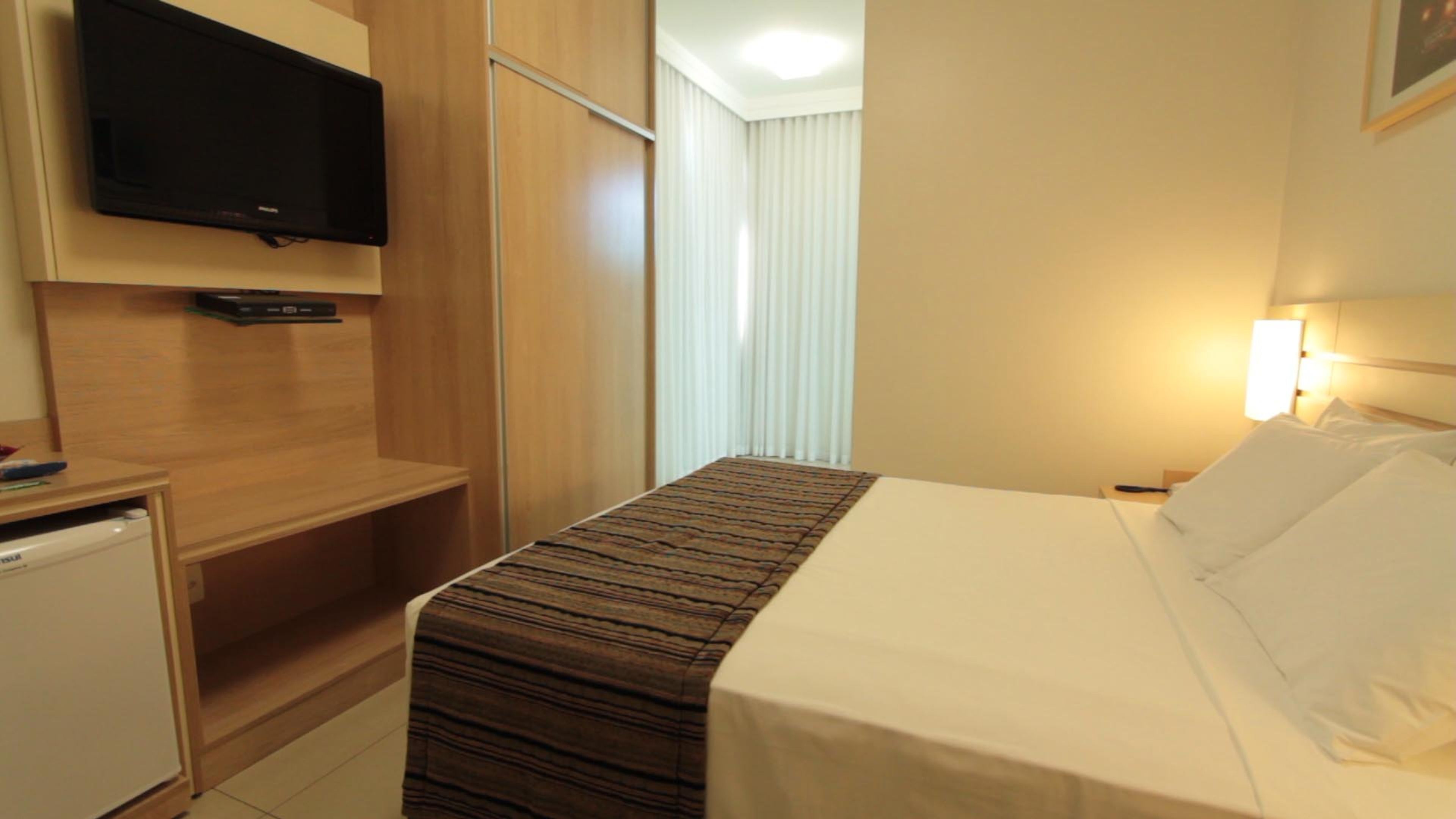 Habitación de hotel con cama, televisor montado en la pared, mesita de noche y ventana con persianas.