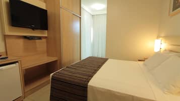 Habitación de hotel con cama, televisor montado en la pared, mesita de noche y ventana con persianas.
