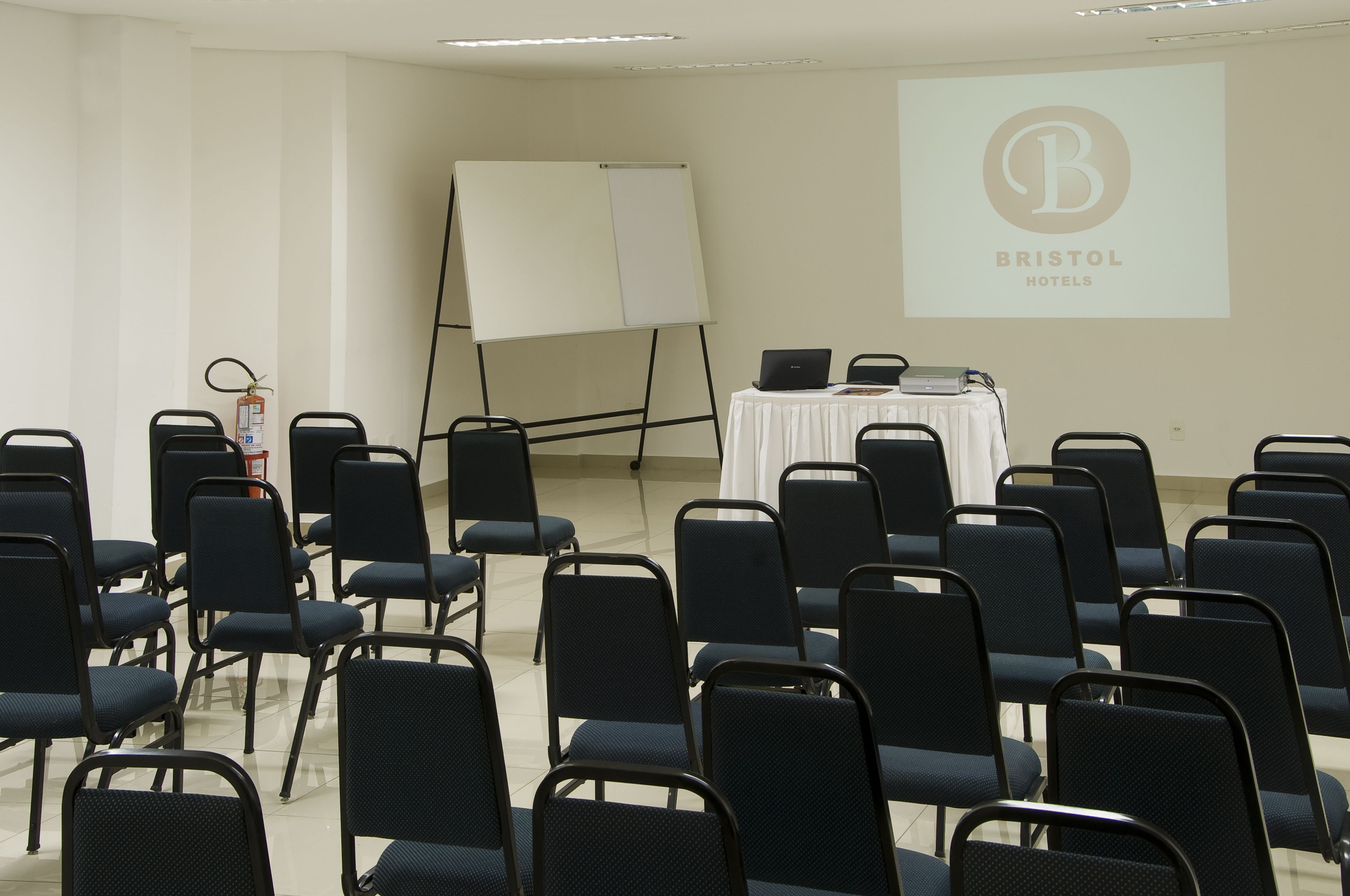 A conference room with rows of chairs, a whiteboard, a laptop on a table, and a projector screen displaying the Bristol Hotels logo.
