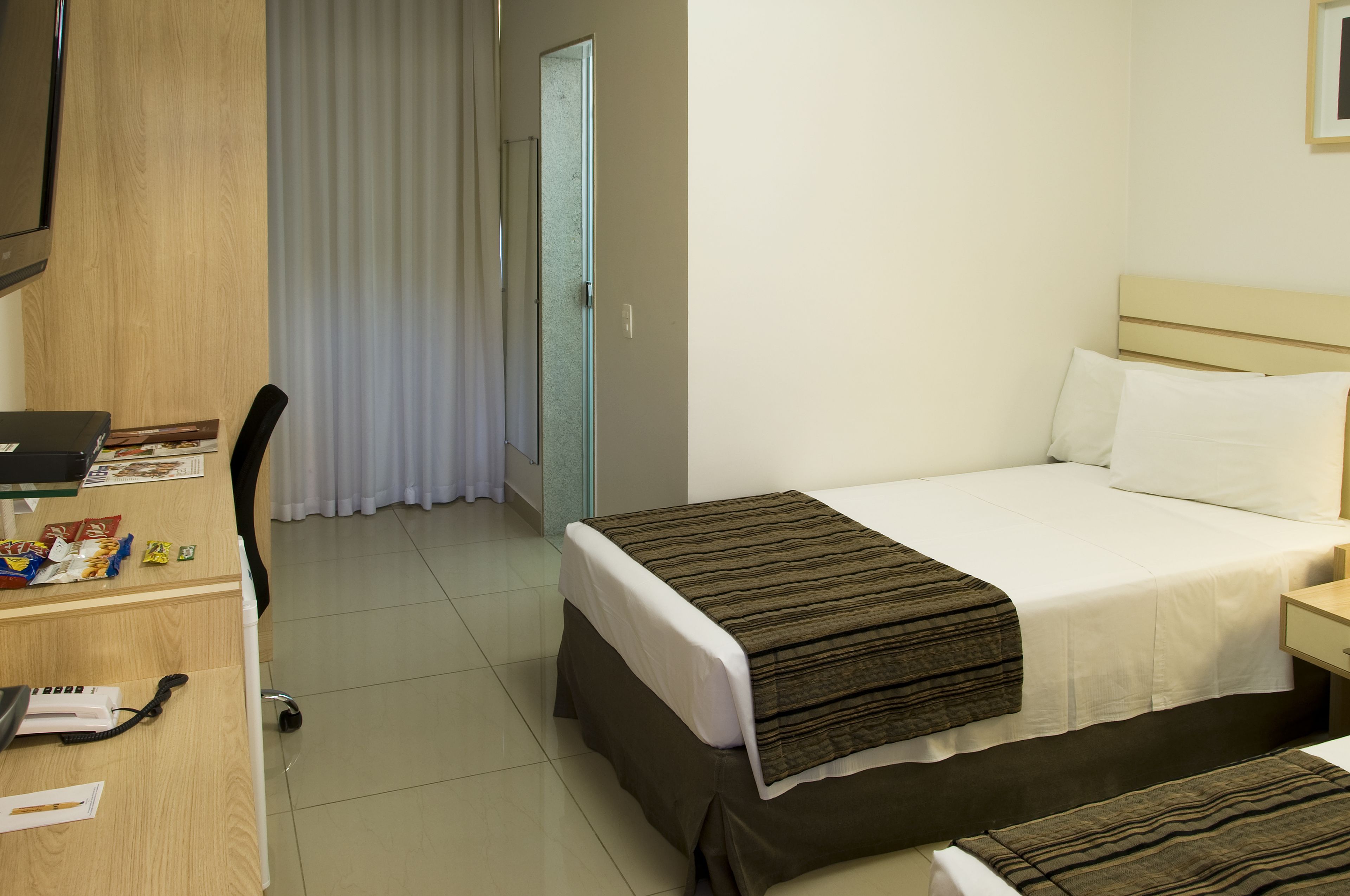 Una habitación de hotel con cama, escritorio con computadora, silla y ventana con cortinas.